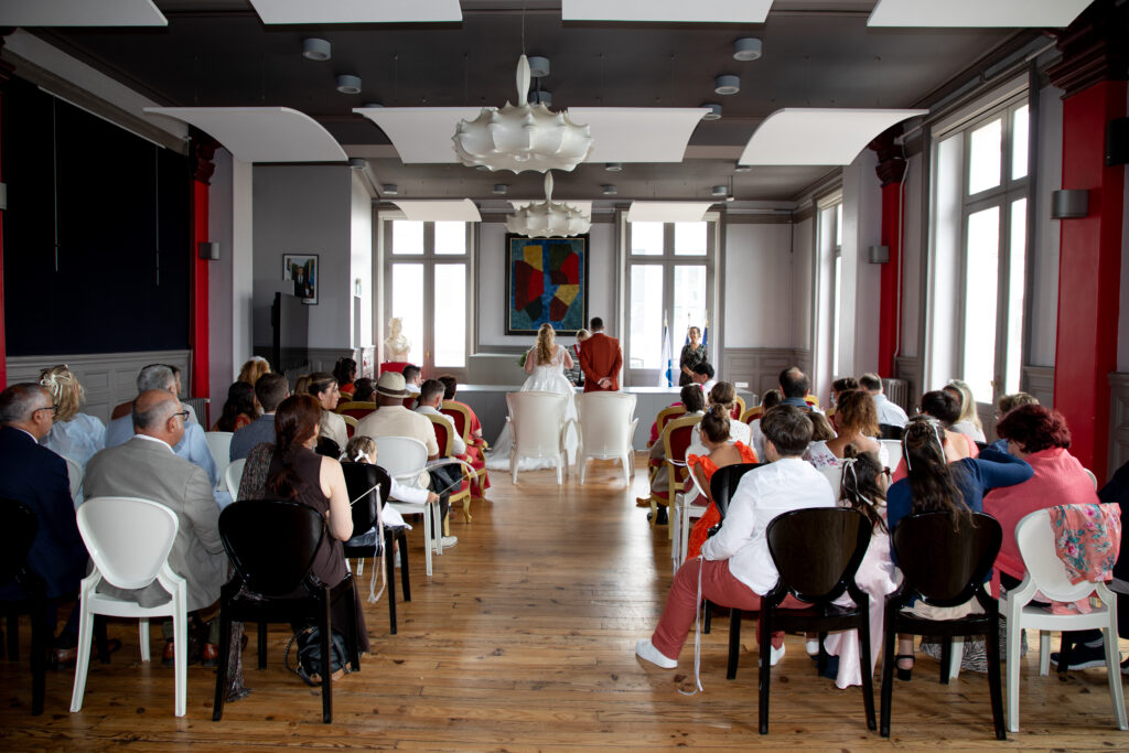 Cérémonie de mariage civil dans une salle de mairie moderne à Bordeaux, vue des mariés et des invités de dos.
