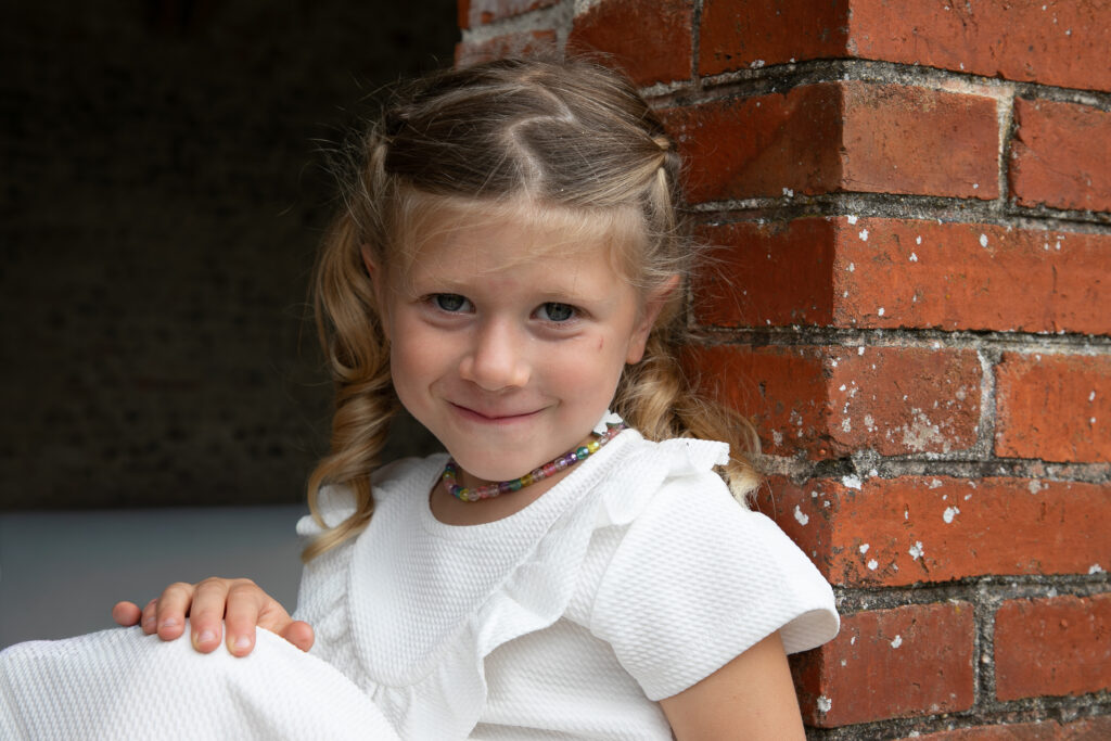 Portrait naturel d'une petite fille d'honneur souriante contre un mur de briques rouges, mariage Bordeaux.